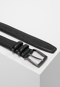 Cinturón de cuero negro con textura suave, hebilla metálica rectangular y múltiples agujeros para ajuste de tamaño. Cuenta con dos lazos de sujeción.