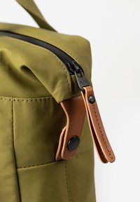 Angolo di una borsa di tessuto verde oliva che mostra una cerniera nera con un manico in pelle beige attaccato da un bottone a scatto nero, su sfondo bianco.