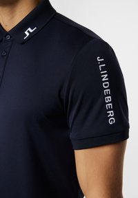 Mörk marinblå polo skjorta med krage, med en logotyp på kragen och "J.LINDEBERG" broderat i vitt på ärmen. Slät textur.