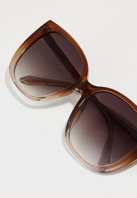 Überdimensionale Sonnenbrillen mit braunen transparenten Rahmen und verlaufsdunklen Gläsern. Glatte Oberfläche mit gewelltem Design und schlanken Bügeln.