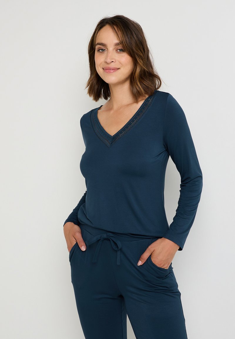 Haut à manches longues bleu marine avec un décolleté en V orné de dentelle. Tissu doux, silhouette épurée, porté avec un pantalon à cordon assorti, poches latérales visibles.