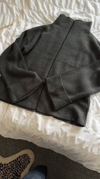 Suéter texturizado gris oscuro con un cuello alto, diseño acanalado y mangas largas, extendido sobre una superficie de tela blanca.