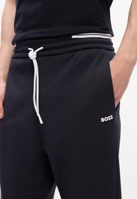 Person iført sorte BOSS sweatpants med hvid snøre og sidelomme, hånd hvilende indeni lommen mod en hvid baggrund.