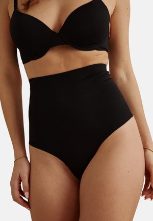 Femme portant un slip gainant noir taille haute et un soutien-gorge noir assorti, debout devant un fond clair uni.