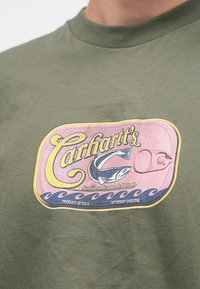 Olívazöld pamut póló színes, retro stílusú halas doboz grafikával, amelyen "Carhartt's" felirat és hullámos mintázat látható.