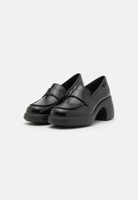 Mustat loafers, joissa on kiiltävä patenttiviimeistely, paksu pyöristetty kärki ja erottuva veistetty korko, jossa on teksturoitu pinta.