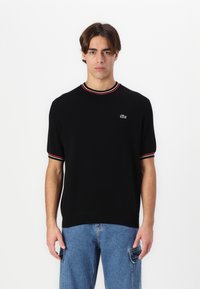Black short-sleeve sweater with red and white striped ribbed collar and cuffs. Features a Lacoste logo on the chest. Smooth texture.