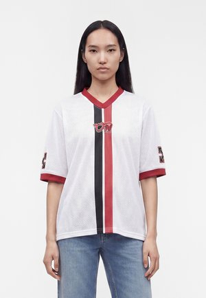 Jeune femme aux longs cheveux noirs porte un t-shirt de sport en maille blanche avec des rayures verticales rouges et noires et un jean bleu, debout devant un fond uni.