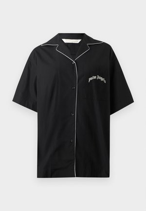 Osoba nosiaca čierne slnečné okuliare, čiernu oversize košeľu s bielym lemovaním a logom, biely crop top, čierne nohavice a strieborné špicaté podpätky.