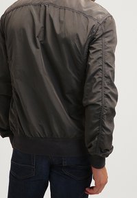 Grå bomberjakke med en glat, skinnende tekstur. Har elastiske manchetter og søm, samt skuldersømme og plisserede ærmer.