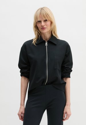 Blonde vrouw met een zwart jack met rits en een borstzak, gecombineerd met een zwarte broek, staand tegen een effen achtergrond.