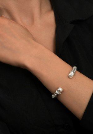 Bracelet en argent avec des accents de crâne détaillés, présentant un design de corde torsadée et des bords lisses, porté sur un poignet contre un fond noir.