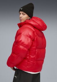 Rode pufferjas met capuchon, met een glanzende, gewatteerde textuur en elastische manchetten. Gecombineerd met een zwarte gebreide beanie.