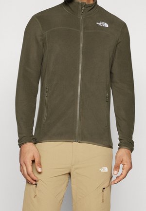 Mężczyzna noszący oliwkowo zieloną polarową kurtkę zapinaną na zamek oraz beżowe spodnie, oba z logo The North Face na piersi i udzie.