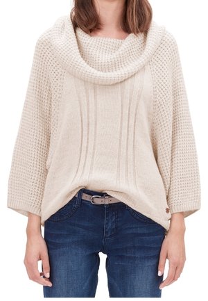 Nainen, joka pitää päällään kermaista paksua kauluksellista neulepuseroa, jossa on leveät hihat, ja tummansiniset farkut, kädet rentona sivuilla.