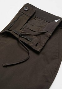 Pantalon marron foncé dézippé avec une ceinture boutonnée et un cordon noir visible sur un fond clair.