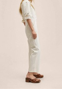 Bílé denimové kšandy s volným střihem, které mají boční kapsy a ohrnuté rukávy, v kombinaci s hnědými chunky klínovými dřeváky se zlatými nýty.