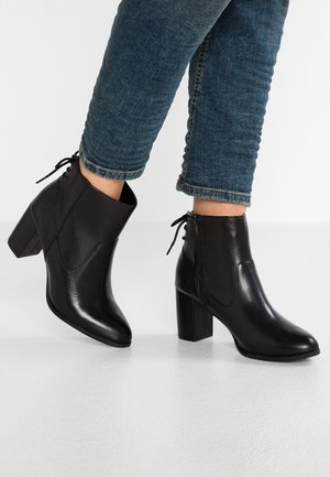 Sorte læder ankelstøvler med spidse tæer, blokhæle og snøredetaljer. Jeans opfoldet over anklen, som eksponerer støvlerne.
