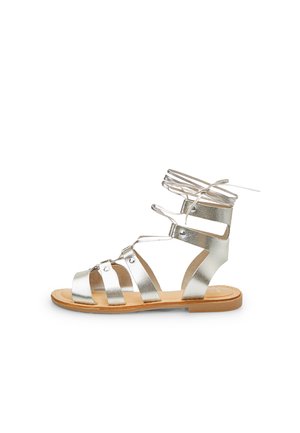 Silver metallic gladiator sandals feature multiple straps, an ankle tie, and a flat tan sole with minimal stitching details.
