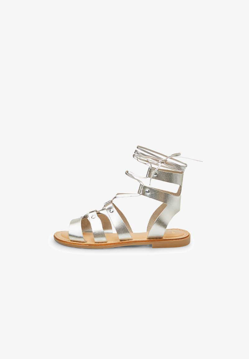 Hopeiset metallinväriset gladiator sandaletit, joissa on useita hihnoja, nilkkasangassa nauha ja litteä ruskea pohja, jossa on vain vähän ompelutietoja.