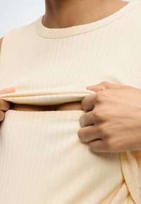 Άτομο σηκώνει ένα καπάκι σε μια κρεμ ριμπ αμάνικη μπλούζα, αποκαλύπτοντας ένα άνοιγμα κάτω από το στήθος για θηλασμό ή άντληση.