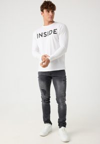 Camisa blanca de manga larga con la palabra "INSIDE" impresa en negro, combinada con jeans oscuros desgastados y zapatillas blancas. Diseño casual y ajustado.