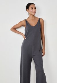 Mono gris con piernas anchas, escote en V profundo y tirantes anudados. Tejido suave con una textura lisa, que presenta una costura en la parte delantera.