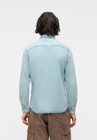 Άτομο με μεσαίου μήκους σκούρα μαλλιά που φοράει ανοιχτό μπλε τζιν πουκάμισο και καφέ φόρμα cargo, εμφανιζόμενο από πίσω με φόντο έναν απλό τοίχο.