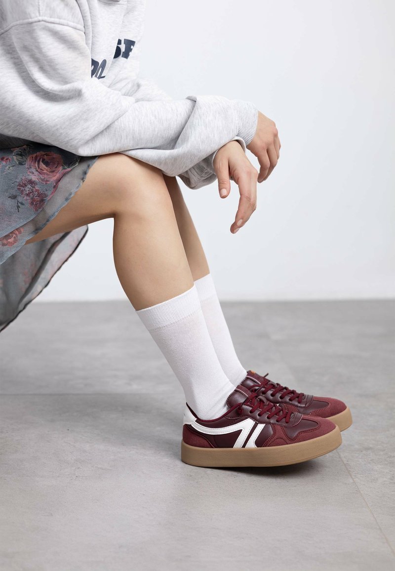 Zapatillas color burdeos con detalles blancos y suela de goma, que presentan un diseño texturizado. Combinadas con calcetines blancos y una falda floral.