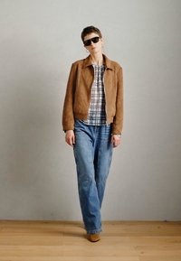 Rjav nubuk jakna preko karirane srajce, v kombinaciji z svetlo modrimi jeans hlačami in rjavimi čevlji. Očala in uhani v obliki obroča dopolnjujejo videz.