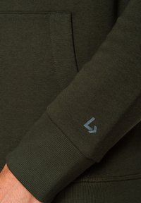 Ciemnozielony materiał bluzy z ribbowanymi ściągaczami i kieszenią. Na rękawie ma małe niebieskie logo. Gładka faktura, zaokrąglony kształt.