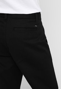 Svarta byxor med en mjuk struktur, slim fit och en enda bakficka med en liten logotyp. Minimalistisk design med subtila detaljer.