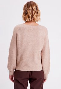 Beige strikket sweater med ribbet tekstur, bred rund udskæring og ballonærmer. Båret med brune bukser, som viser detaljerne på ryggen.