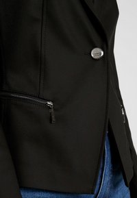 Blazer noir avec un bouton argenté marqué et des poches zippées porté sur un jean en denim bleu.