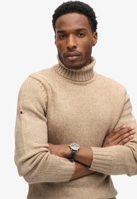 Beżowy sweter z golfem wykonany z dzianiny z prążkowanymi akcentami. Posiada małe logo na rękawie. Prosty i dopasowany krój.
