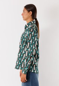 Camicia a maniche lunghe in un tessuto leggero e liscio con un motivo geometrico di forme nere, blu e arancioni su uno sfondo bianco.