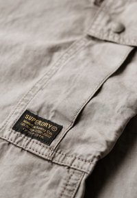 Hellgrau Stoff mit glatter Textur, versehen mit einem schwarzen Etikett mit goldener Schrift, auf dem "SUPERDRY" steht. Mit Knopfdetail am Kragen.