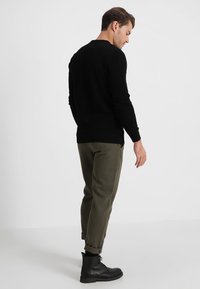 Zwarte gestructureerde trui met een geribbelde zoom, gecombineerd met olijfgroene tapered pants en zwarte enkellaarsjes. Gezien vanaf de achterkant.