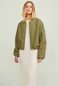 Jaqueta bomber verde oliva com fecho à frente, punhos canelados e um bolso com tampa na manga esquerda, usada sobre um vestido longo bege.