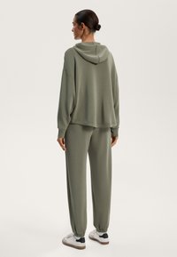 Natoľko veľká olivovo zelená mikina s kapucňou, v kombinácii so zladenými zúženými teplákmi, s elastickými manžetami a vo voľnom strihu.