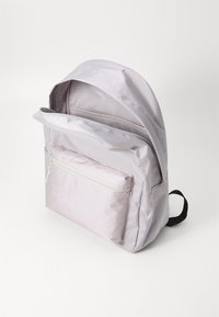 Mochila lilás clara feita de tecido durável. Apresenta um bolso frontal com fecho e alças ajustáveis. Textura suave com uma marcação discreta.