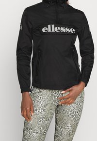 Sort vindjakke med en fremtrædende grå "ellesse"-logo, lynlåslommer og elastiske manchetter, kombineret med mønstrede leggings.