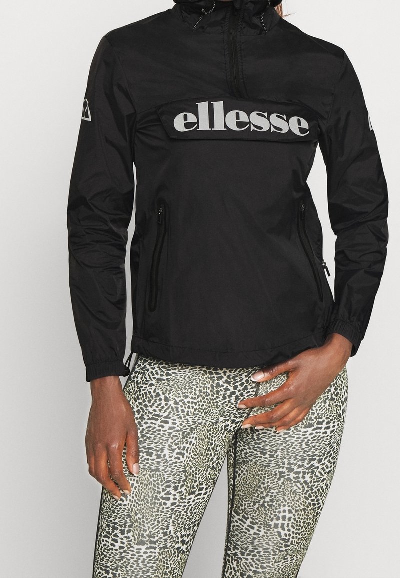 Sort vindjakke med en fremtrædende grå "ellesse"-logo, lynlåslommer og elastiske manchetter, kombineret med mønstrede leggings.