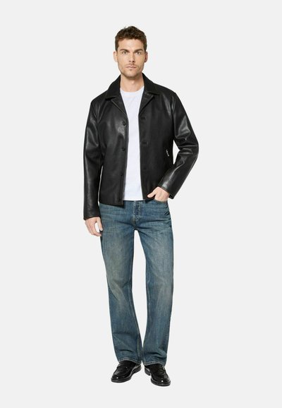 Man wearing black leather jacket, white t-shirt, blue jeans, and black shoes, standing with one hand in pocket against white background.