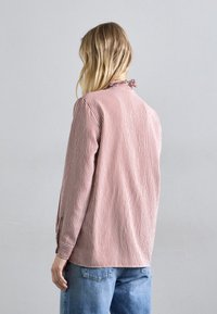 Camicia a maniche lunghe con strisce verticali rosse e bianche, tessuto morbido, colletto arrotondato e polsini con bottoni, abbinata a jeans azzurri chiari.