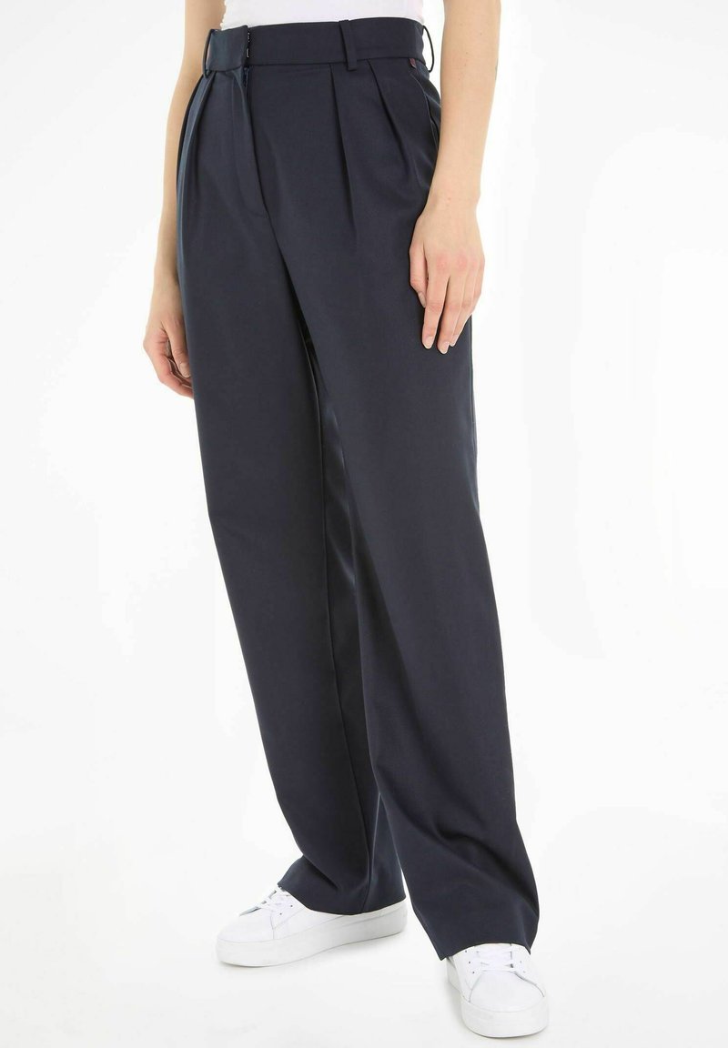 Pantalons taille haute bleu marine avec des plis, confectionnés en tissu lisse. Présente une coupe ajustée et des poches latérales ; portés avec des baskets blanches.