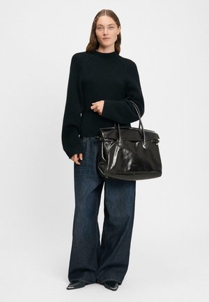Czarna skórzana torba na ramię o strukturalnym kształcie, z dwoma uchwytami u góry i błyszczącym wykończeniem. Nosi z ciemnym swetrem i szerokimi spodniami dżinsowymi.