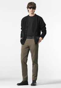 Jonge man in een zwarte cardigan, olijfgroene broek, zwarte riem, zwarte laarzen en zonnebril, die met handen in de zakken staat op een witte achtergrond.