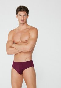 Bordeaux trenýrky s bezšvovým elastickým pasem, přiléhavým střihem a hladkým materiálem. Model s překříženýma rukama, který předvádí detailní záběr na trenýrky.