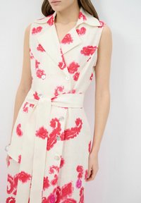 Vestito senza maniche con stampa floreale in modelli rossi e rosa su sfondo bianco. Presenta un design avvolgente, dettagli con bottoni e una cintura da annodare.